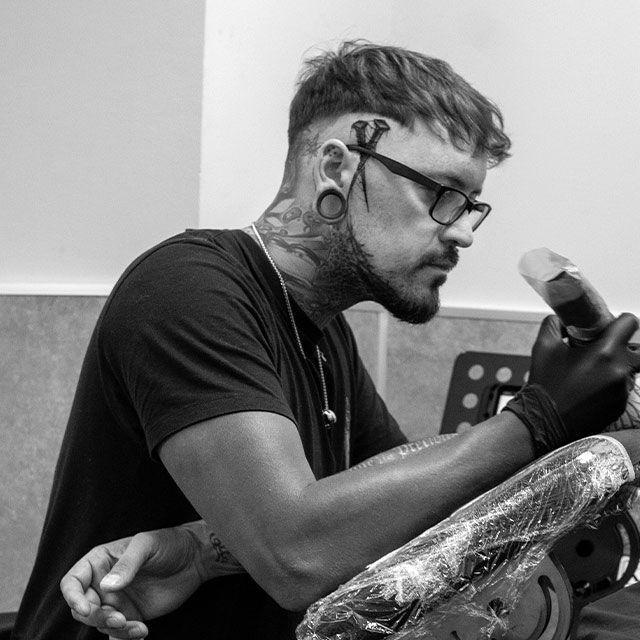 Tattoo artist wearing gloves works on a client’s arm in a black-and-white studio scene